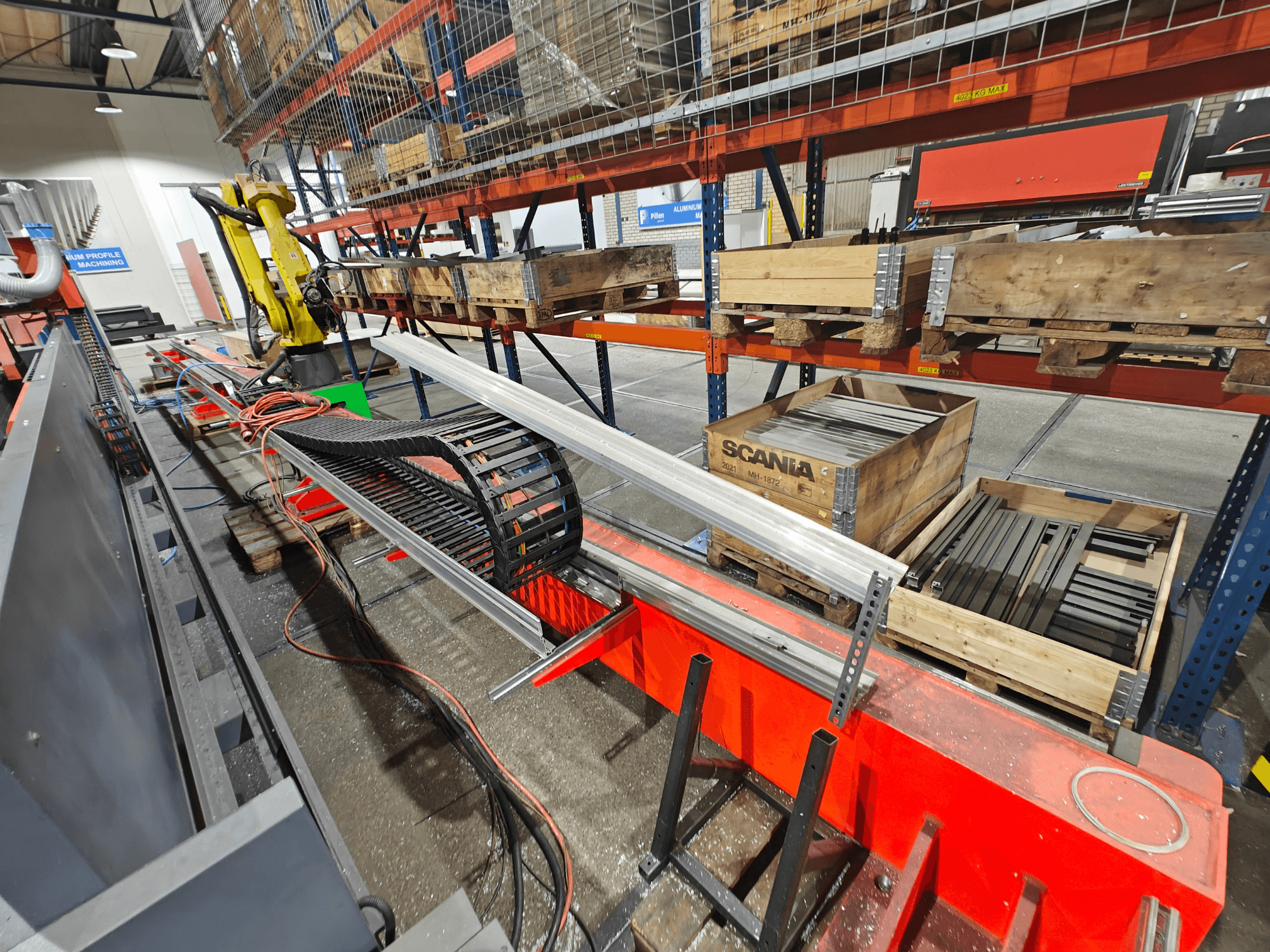 Robotic arm on workstation, orange and black cables visible, with Scania crates and aluminum profiles in a factory setting.