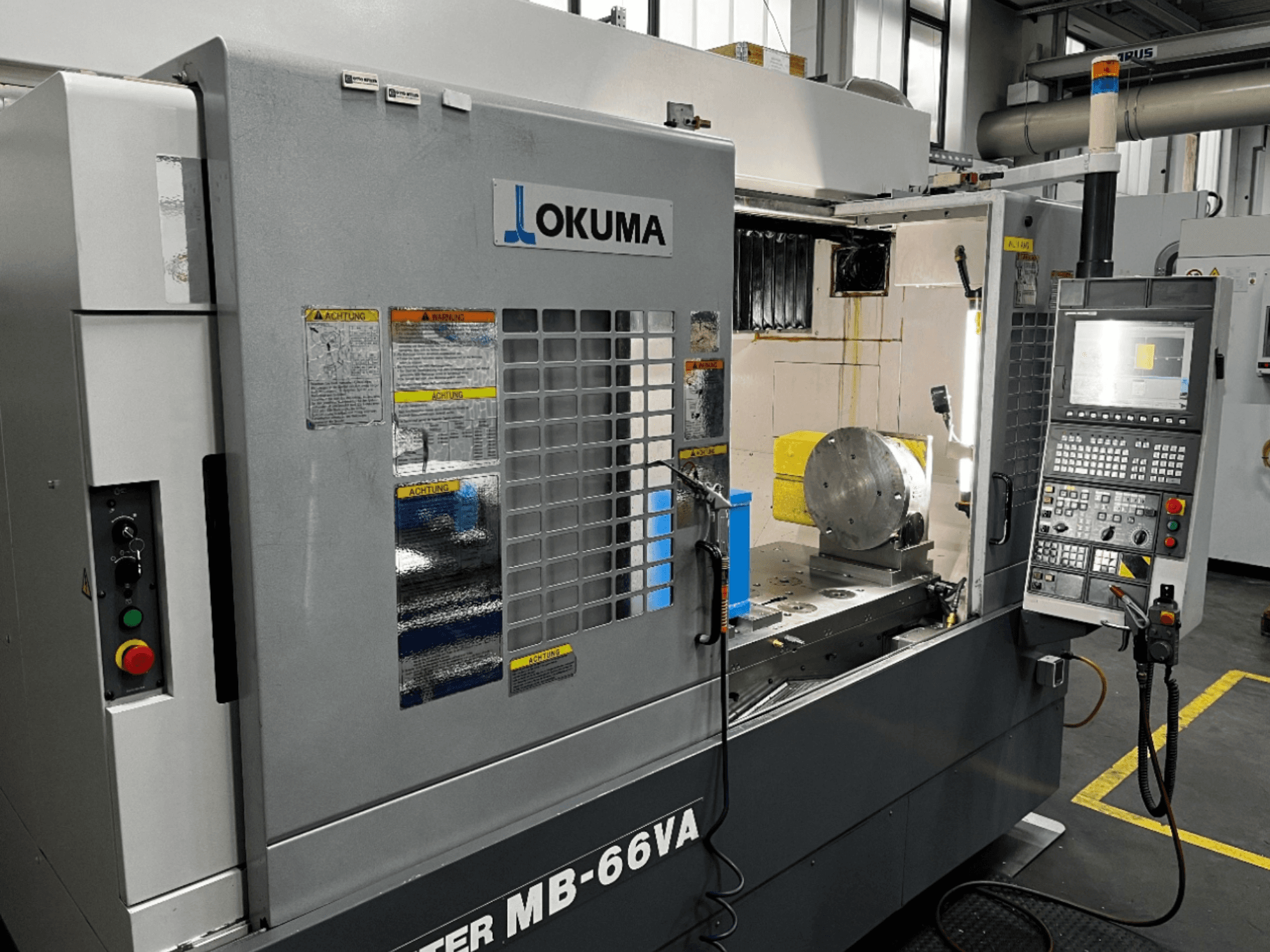 Okuma MB66-VA Vertical Machining Centre front view showing control panel, workpiece, and machine interior with safety labels.