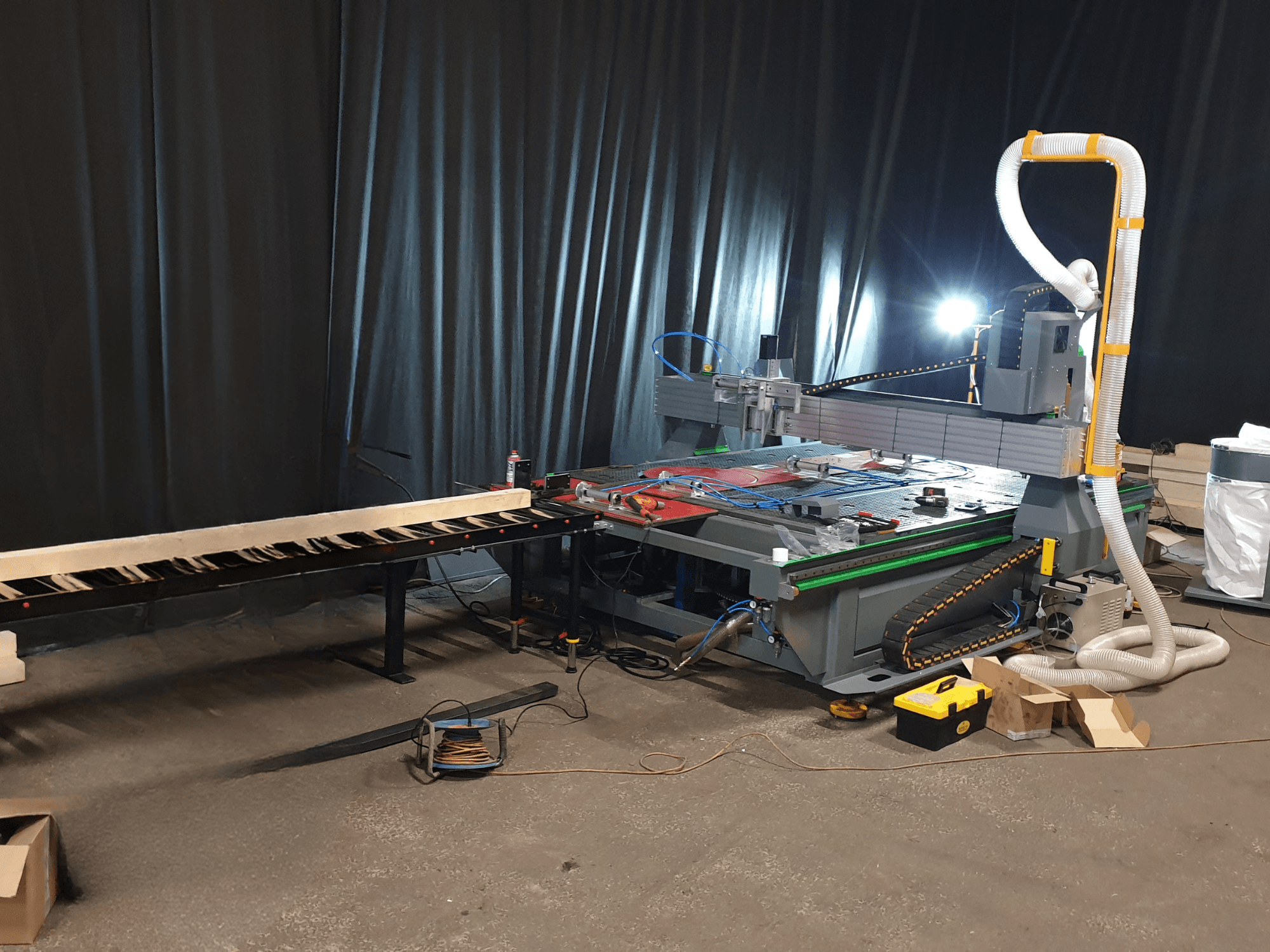 CNC machine with a side conveyor, tools on the table, and duct for dust collection. Black backdrop, front view.