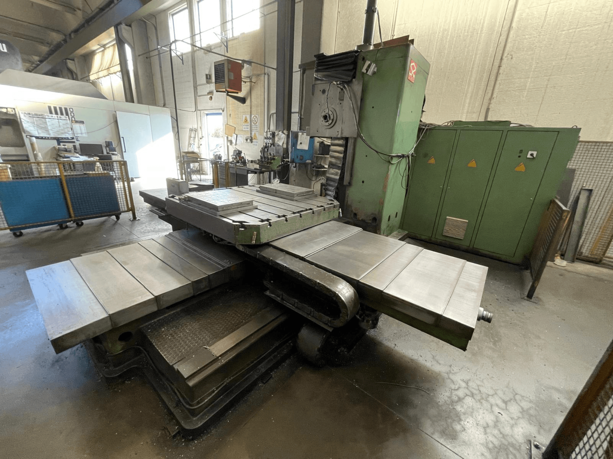 Large industrial milling machine with a green body, side view, featuring multiple flat surfaces and storage cabinets in the background.