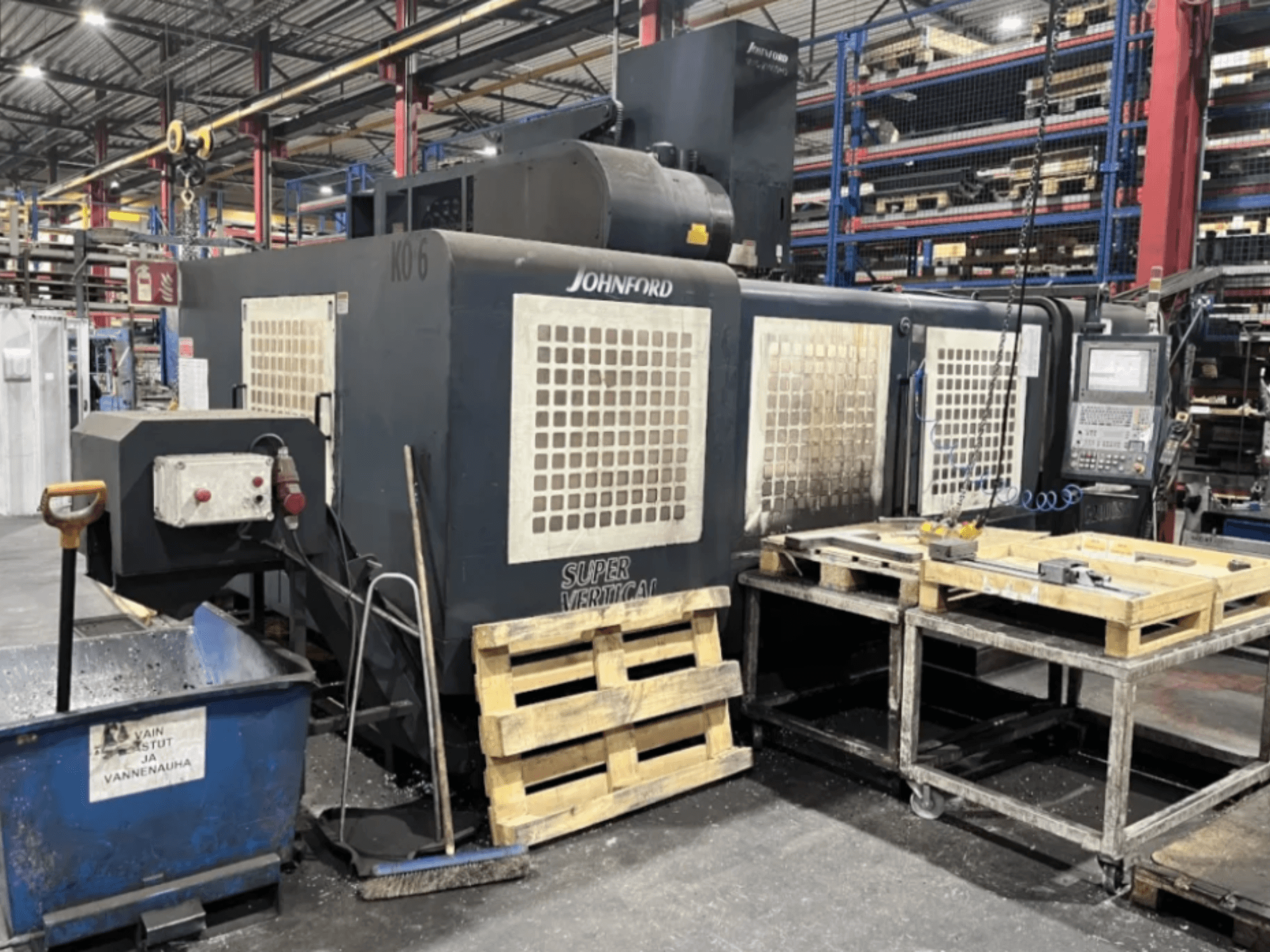 Johnford Super Vertical milling machine in a factory, side view with control panel, pallets, and cleaning equipment nearby.
