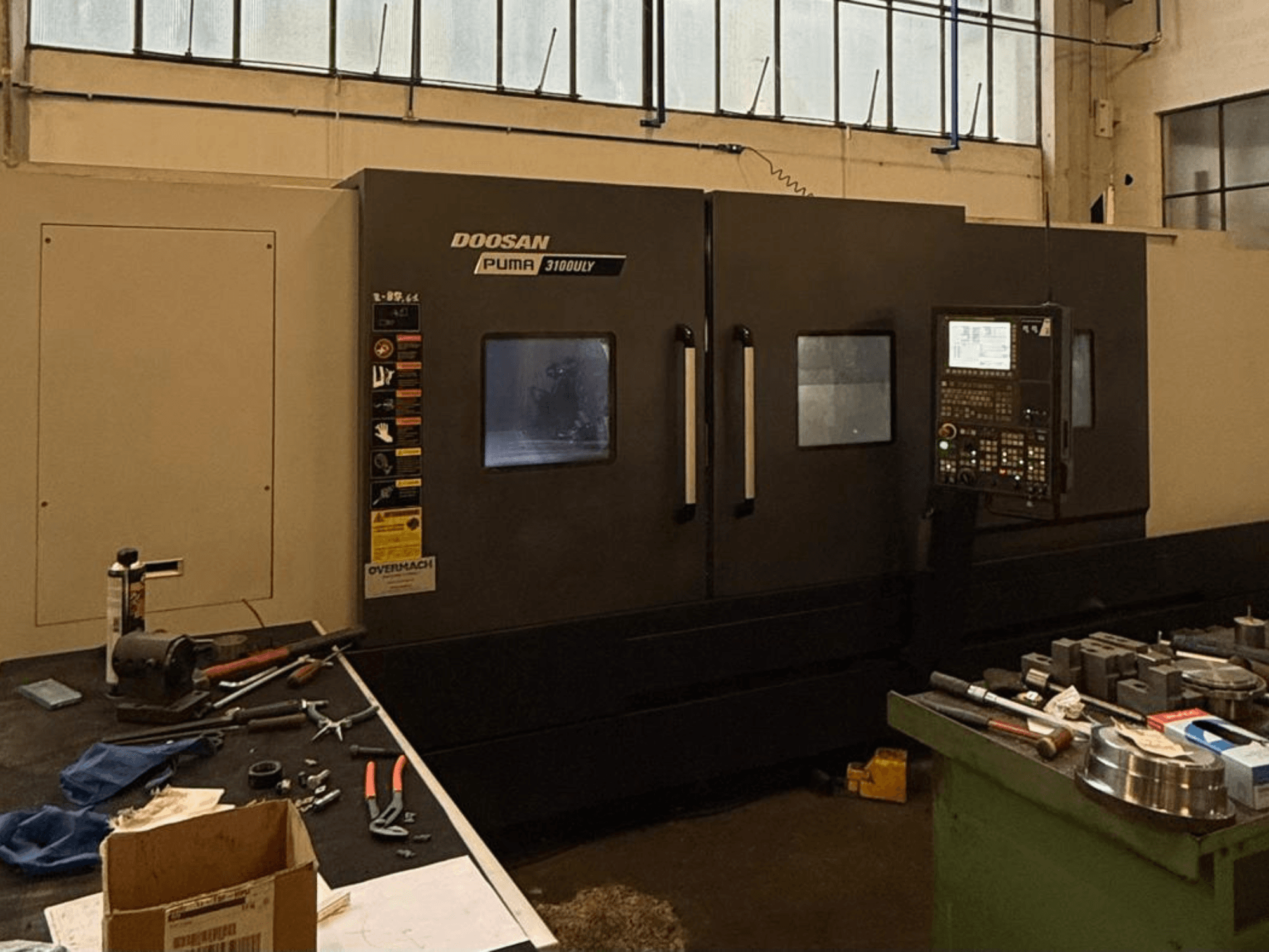 Doosan Puma 3100LY CNC lathe in a workshop, side view, with control panel and tools visible on the workbench.