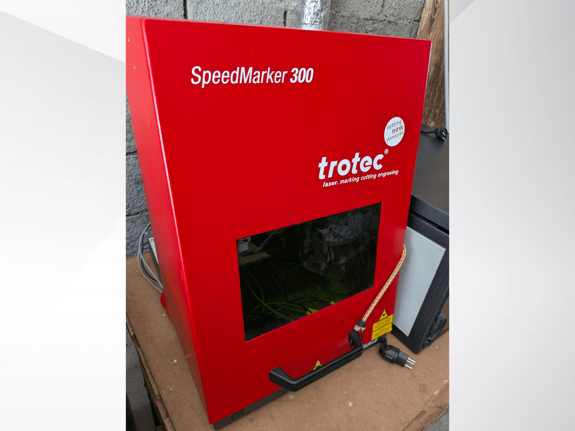 Red Trotec SpeedMarker 300 laser marking machine in front view, featuring a large viewing window and power connection.