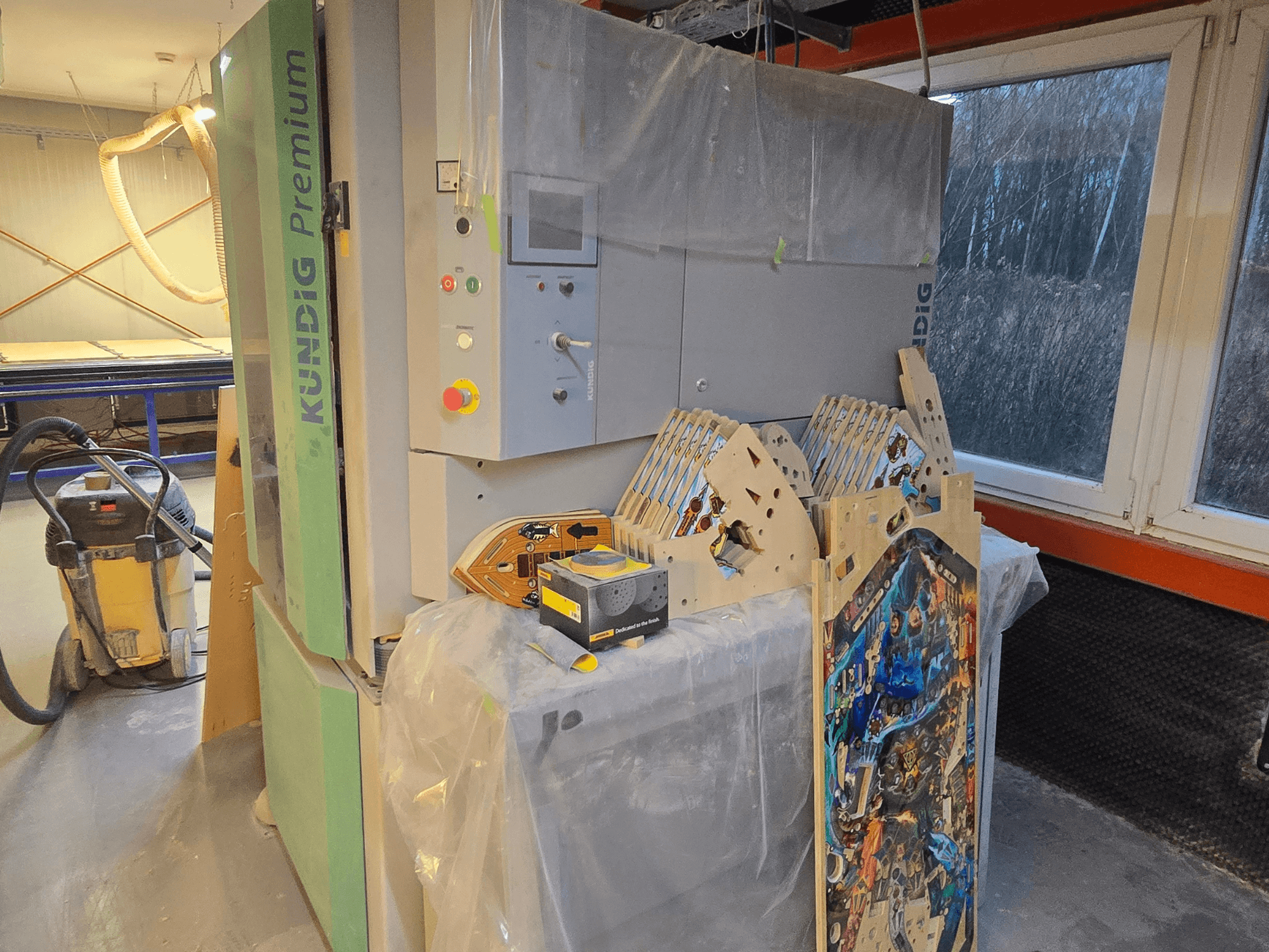 Kundig Premium-1 1100-C belt sander in workshop, showing control panel and wood pieces, side view, with dust vacuum in background.