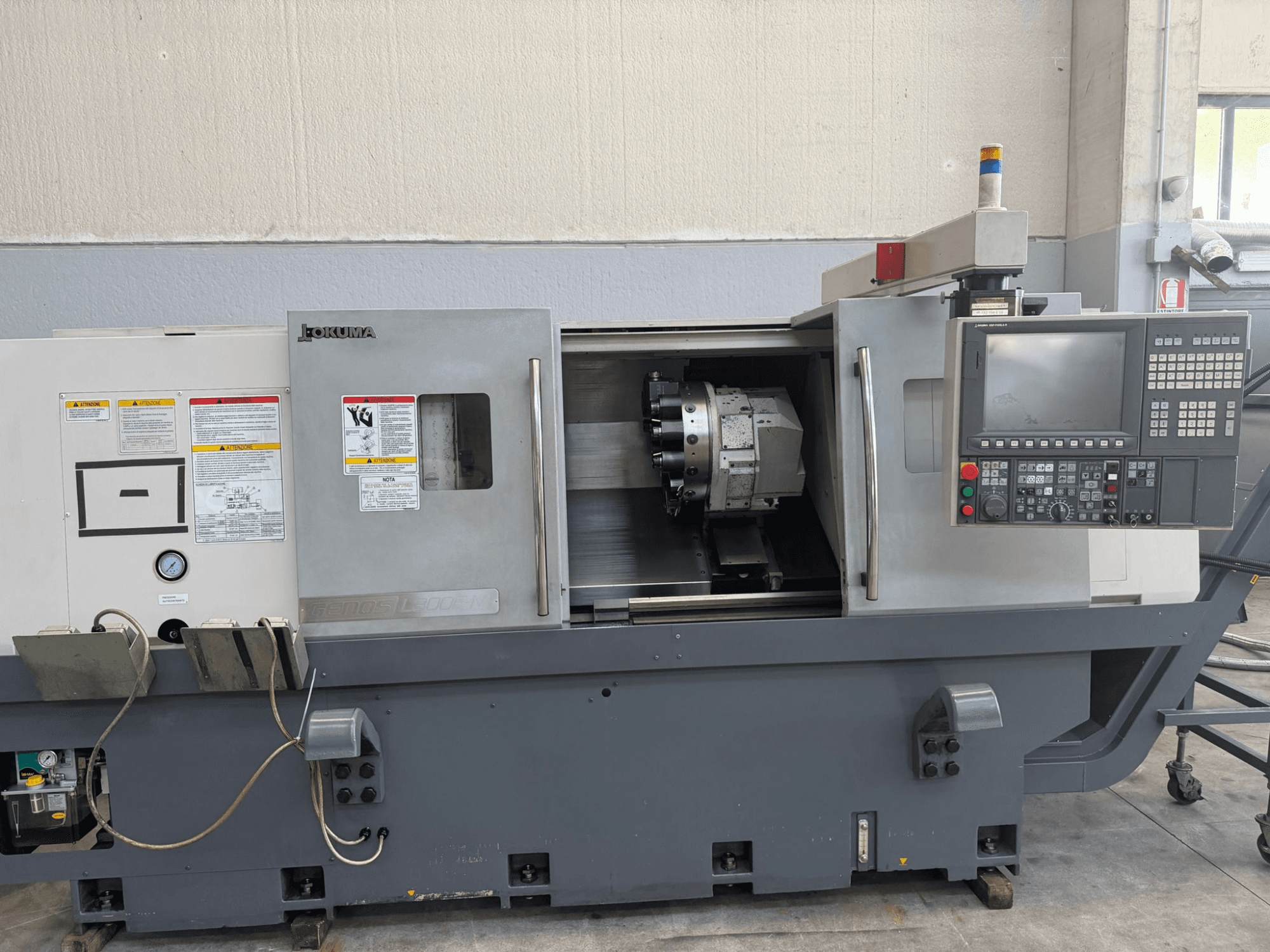 Okuma CNC lathe machine, front view, showing control panel, cutting tool holder, and safety warnings on the side.