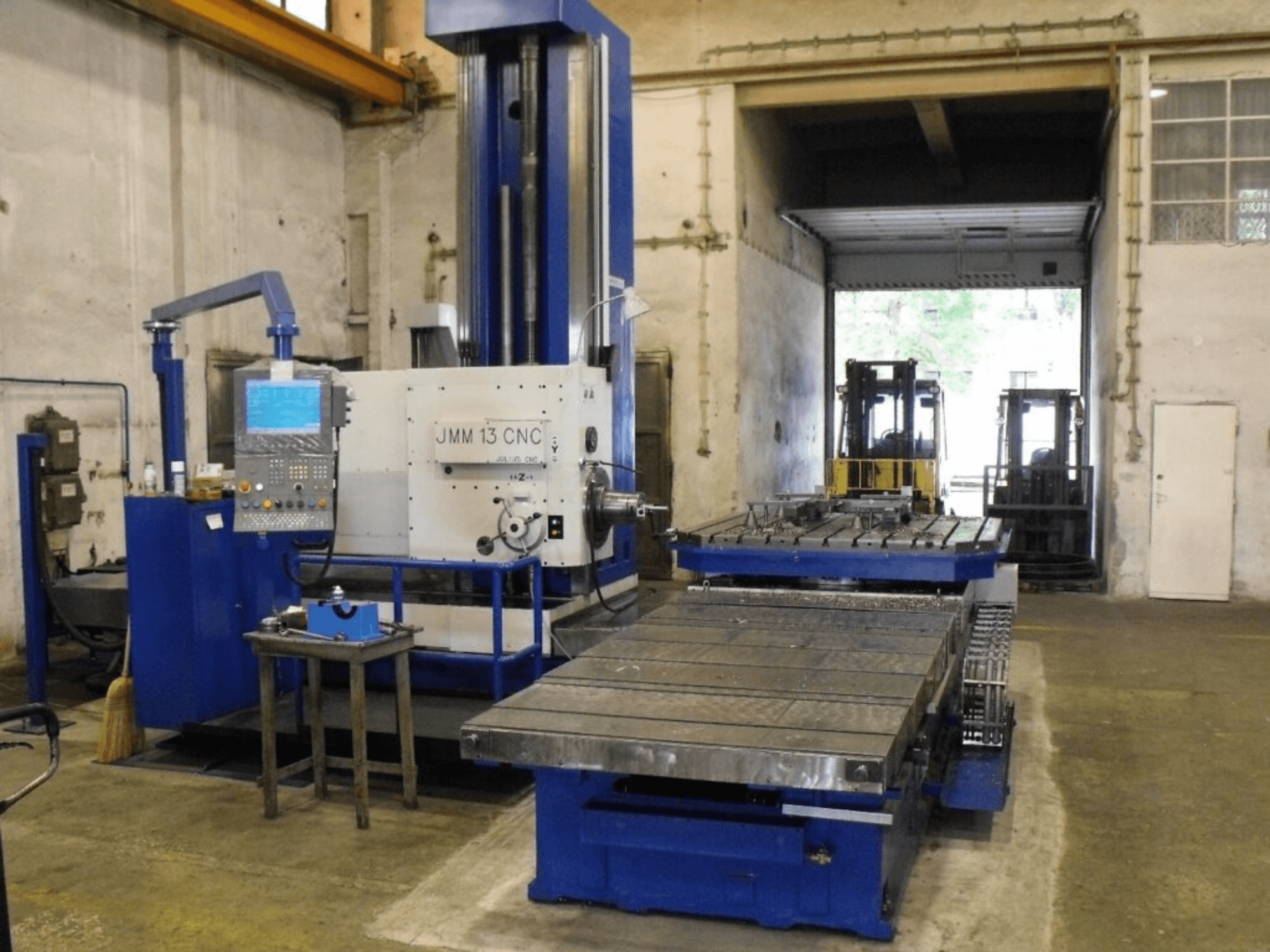 JMM 13 CNC milling machine in a workshop, front view, features a control panel, large work table, and industrial backdrop.