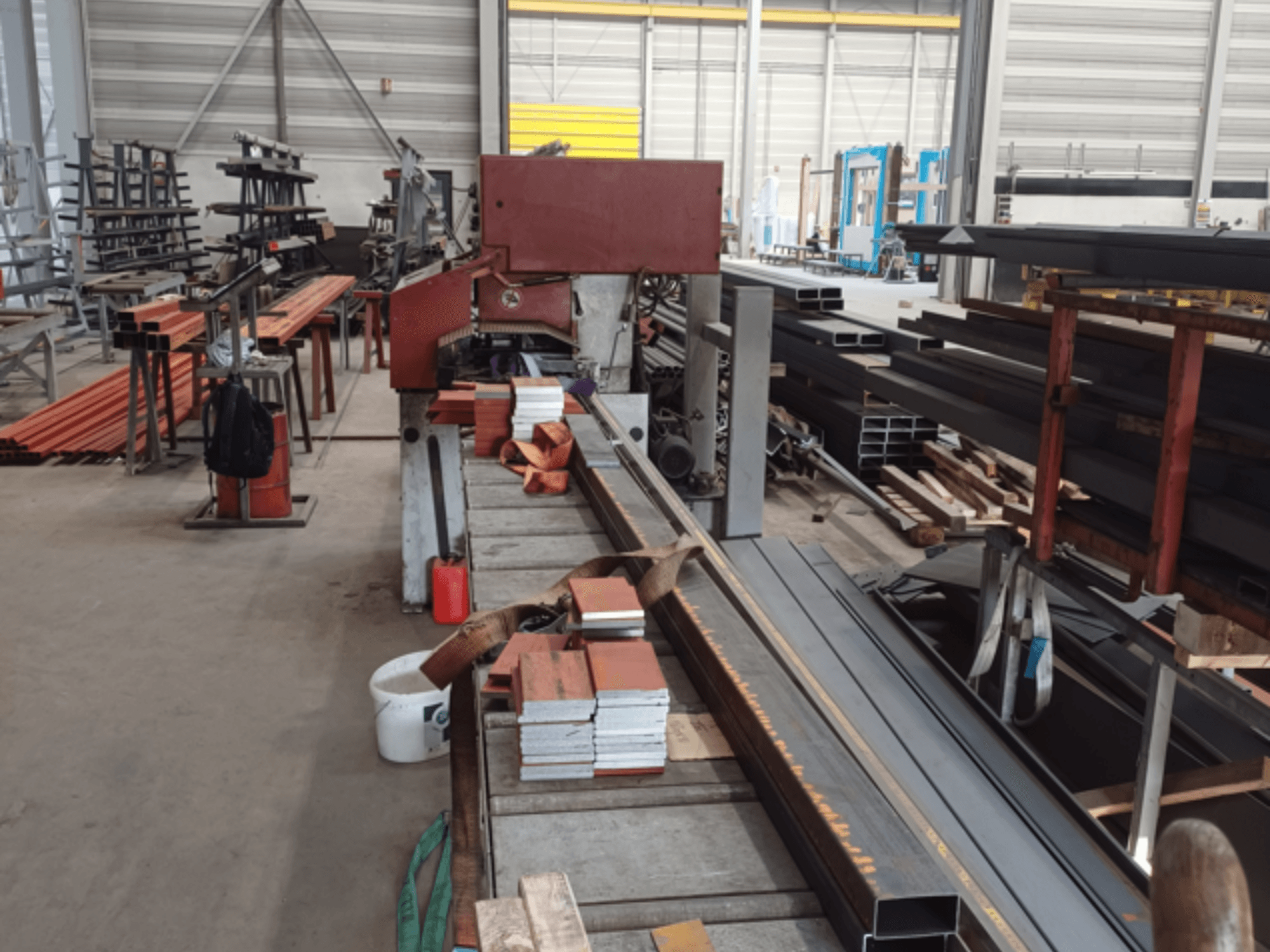 Manufacturing workshop featuring various metal profiles, with a red saw machine in the foreground and stacked materials.