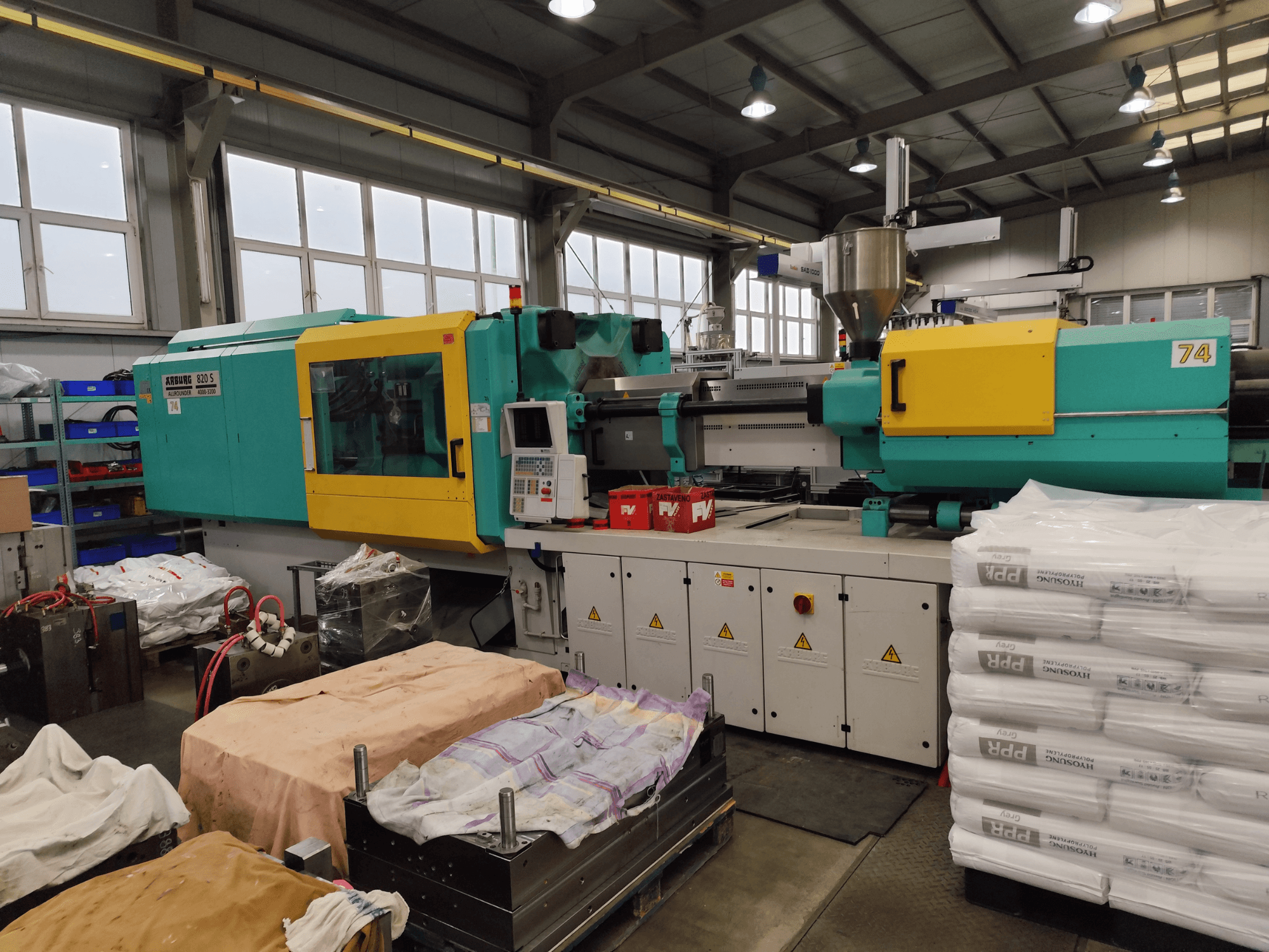 Injection molding machine by Arburg, front view, teal and yellow exterior, with tooling and materials nearby.