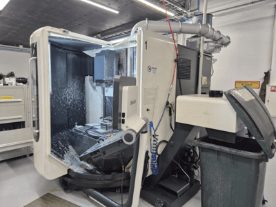 Open DMG milling machine with coolant spray, displaying metal shavings and tool controls, viewed from the front.