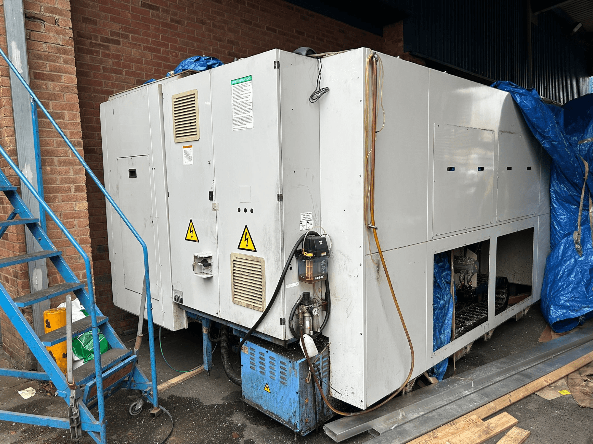 Side view of a large industrial chiller, brand and model unclear, showing electrical connections, warning signs, and blue tarp cover.