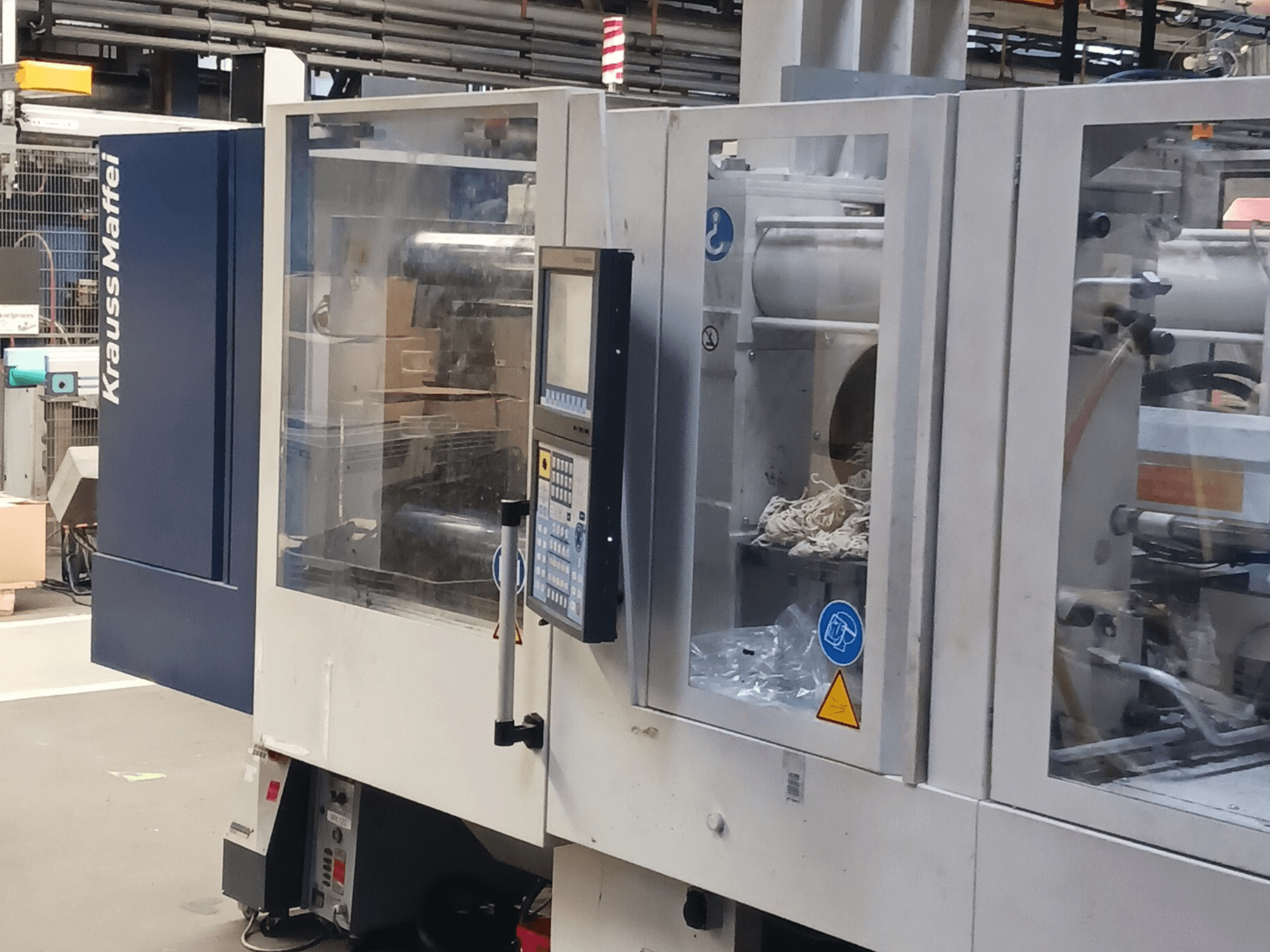KraussMaffei injection molding machine, side view, showing control panel, machinery, and packaging materials in background.