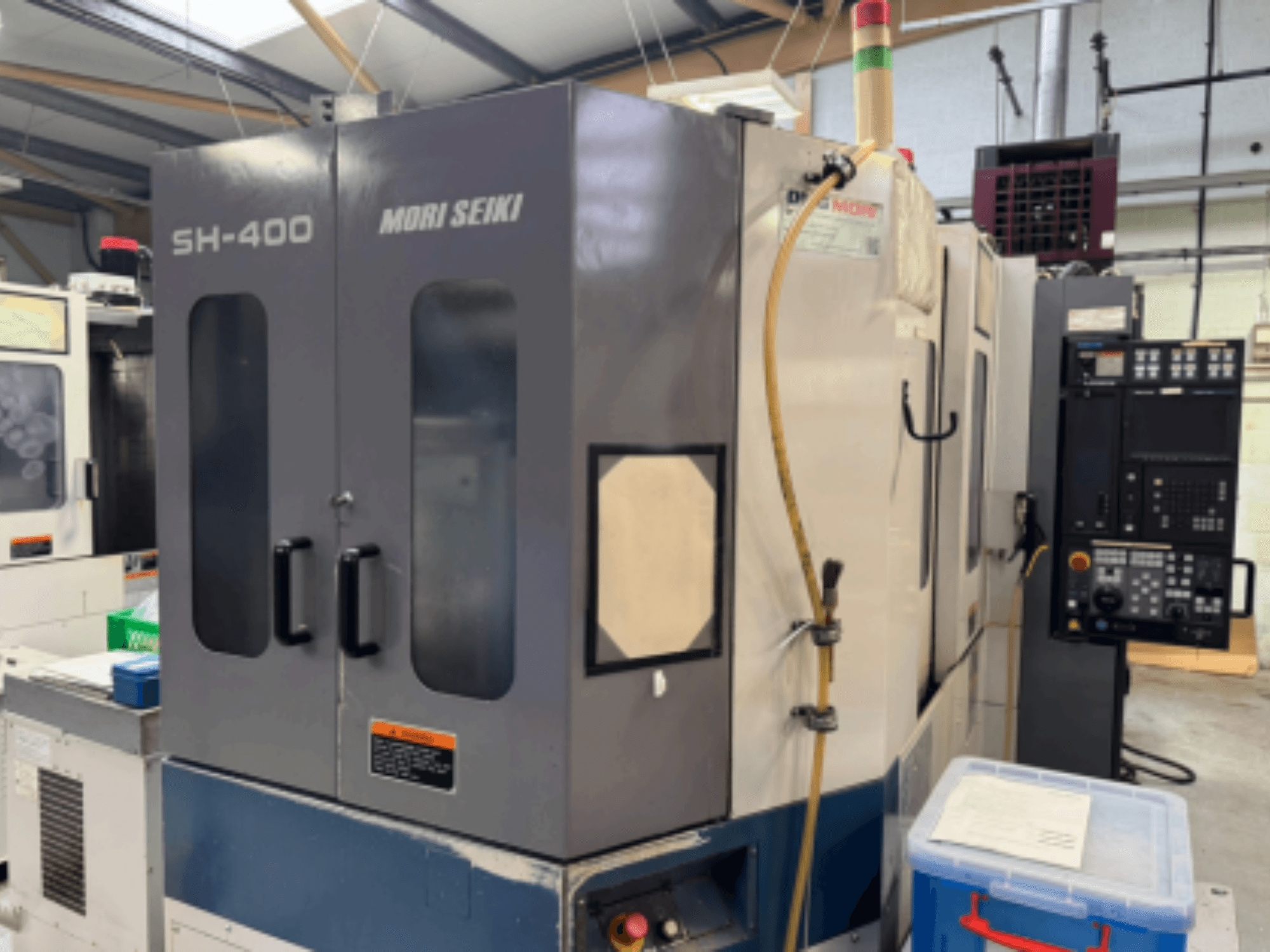Mori Seiki SH-400 CNC machine, front view, featuring dual doors, control panel, and attachment hose. Blue storage container nearby.