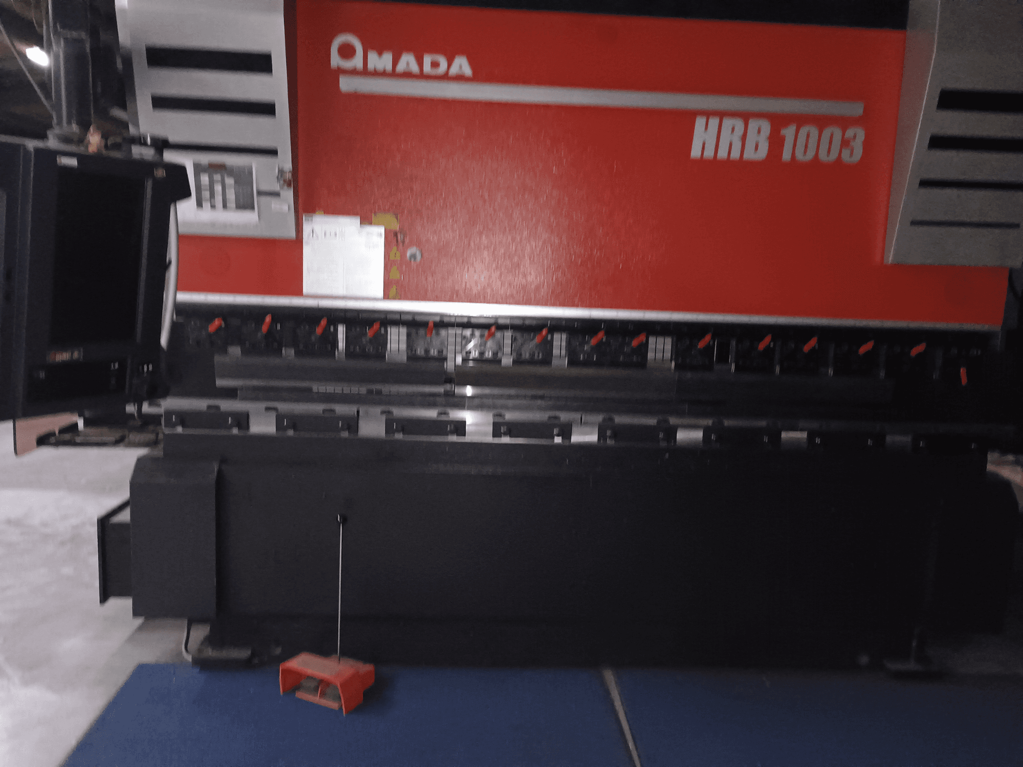 Amada HRB 1003 press brake, front view; features control panel with red knobs and black casing, set in an industrial environment.