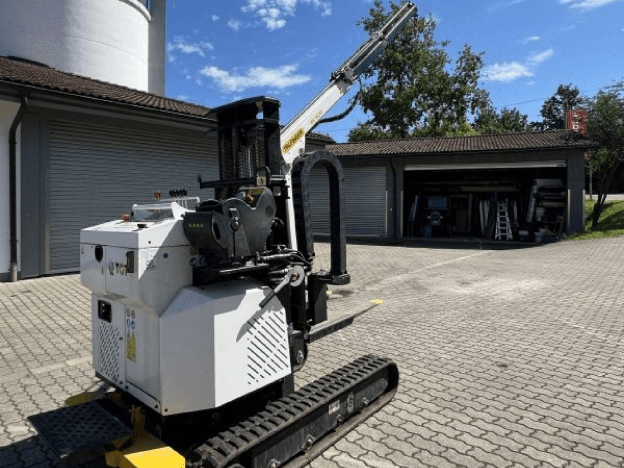 TGI construction mini crane, side view, showcasing tracks and mounted boom, parked on a brick surface by a garage.