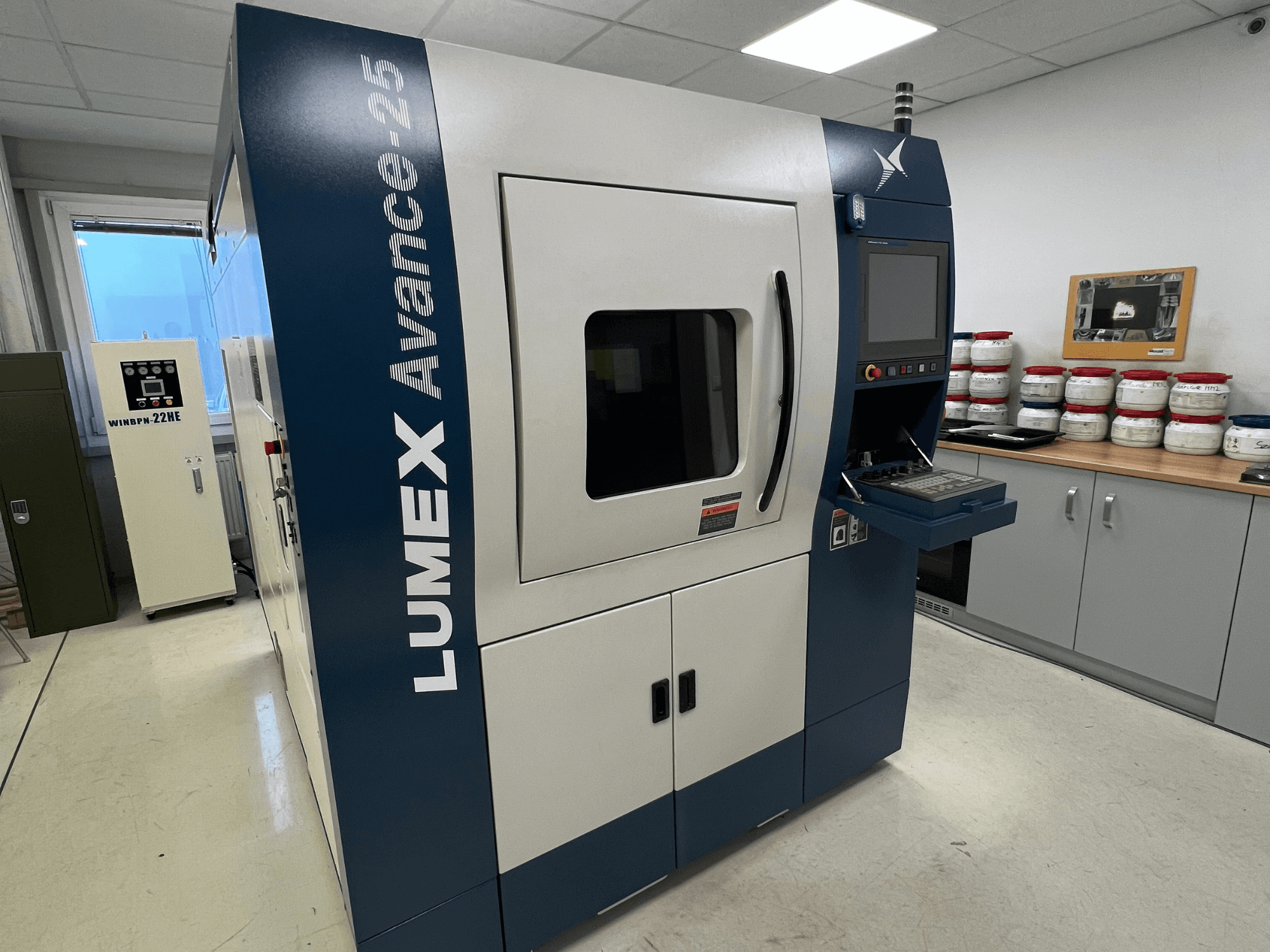LUMEX Avance-25 3D printer in a lab setting, front view. Control panel with screen and buttons visible, equipment in background.