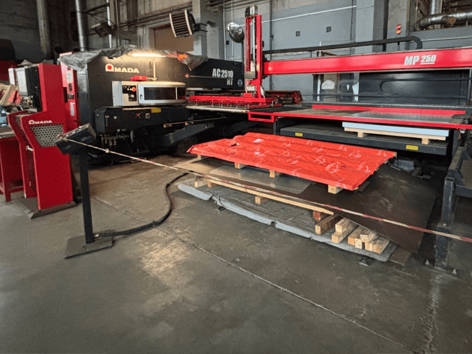 Amada AC2510 NT laser cutter in factory setting, front view, heavy machinery with metal sheets and pallets on the floor.