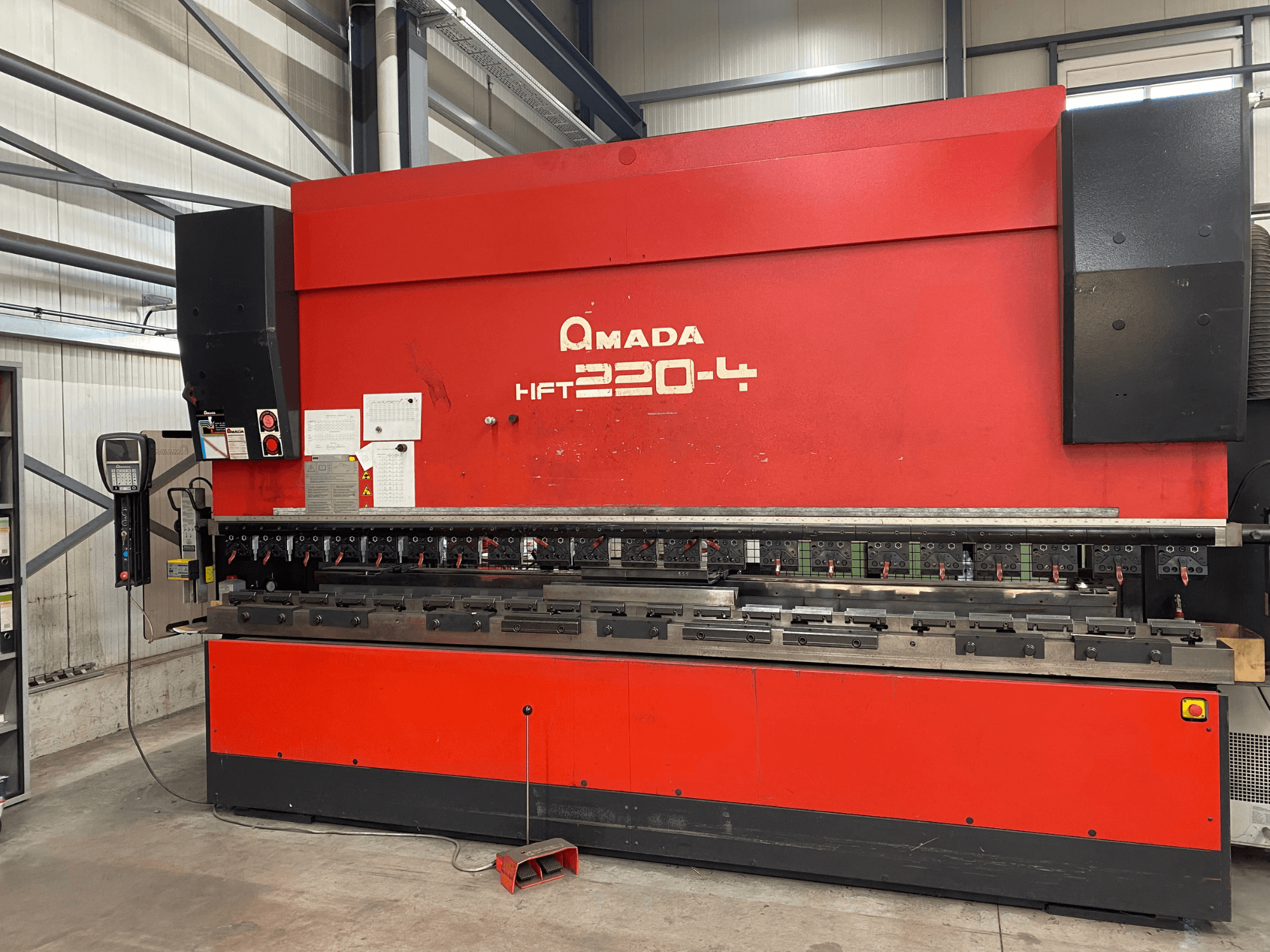 Red AMADA HFT 220-4 press brake in a factory setting, front view showing control panel and bending tools on the work surface.