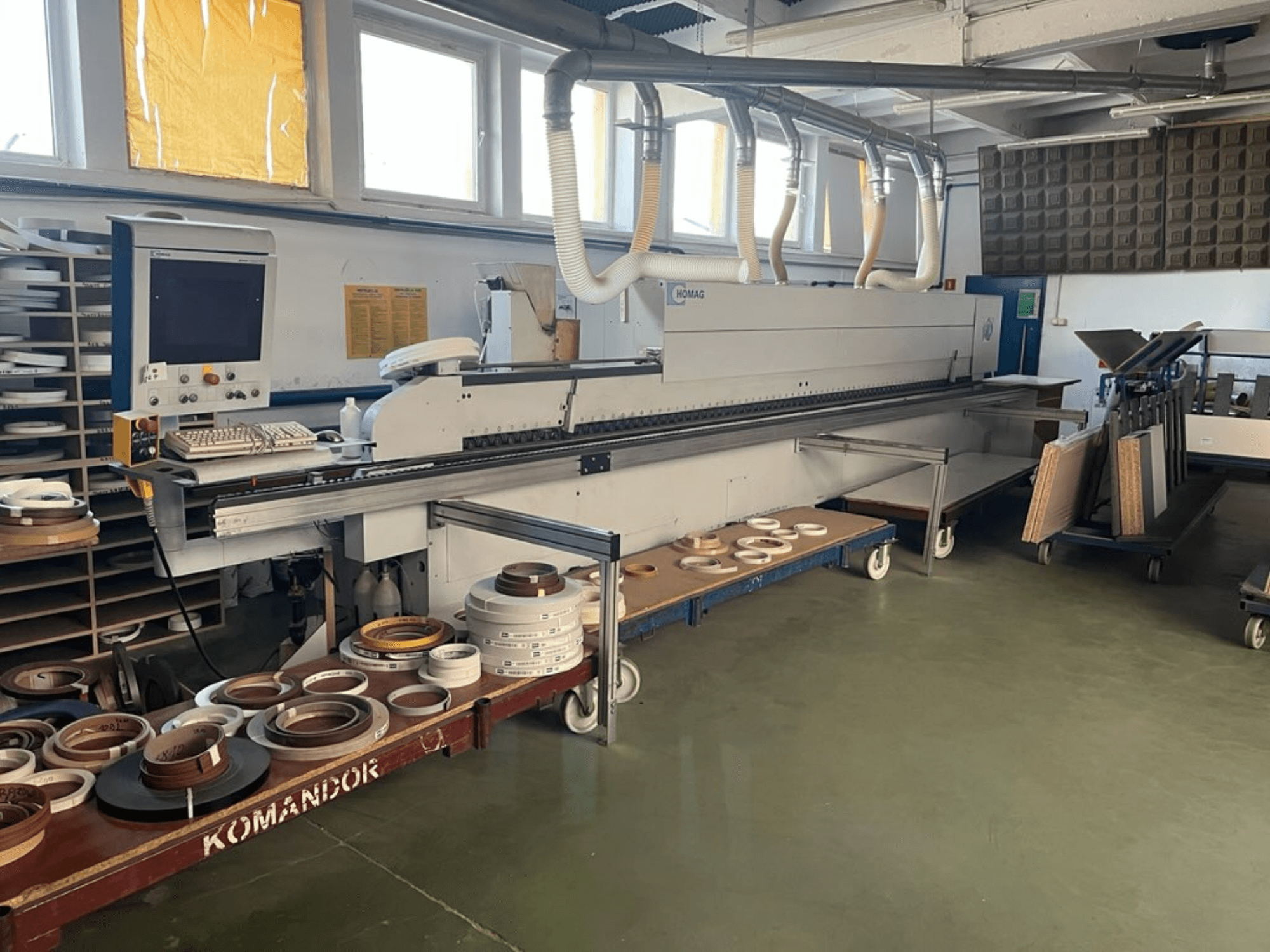 HOMAG edge banding machine in a workshop, featuring controls and various edge banding materials on a blue cart. Side view.