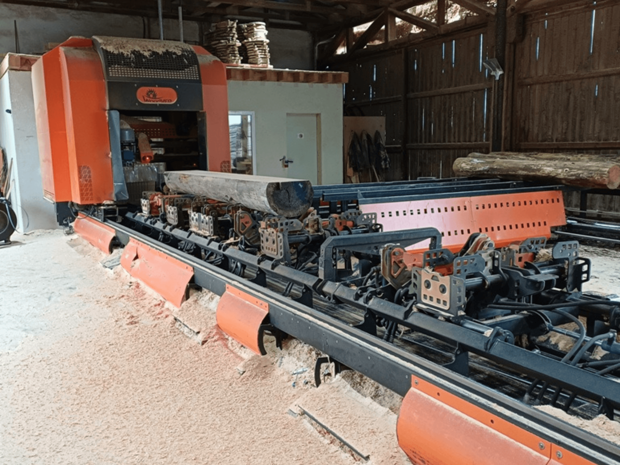 Orange lumber milling machine with a log positioned on the conveyor, viewed from the front in a workshop setting.
