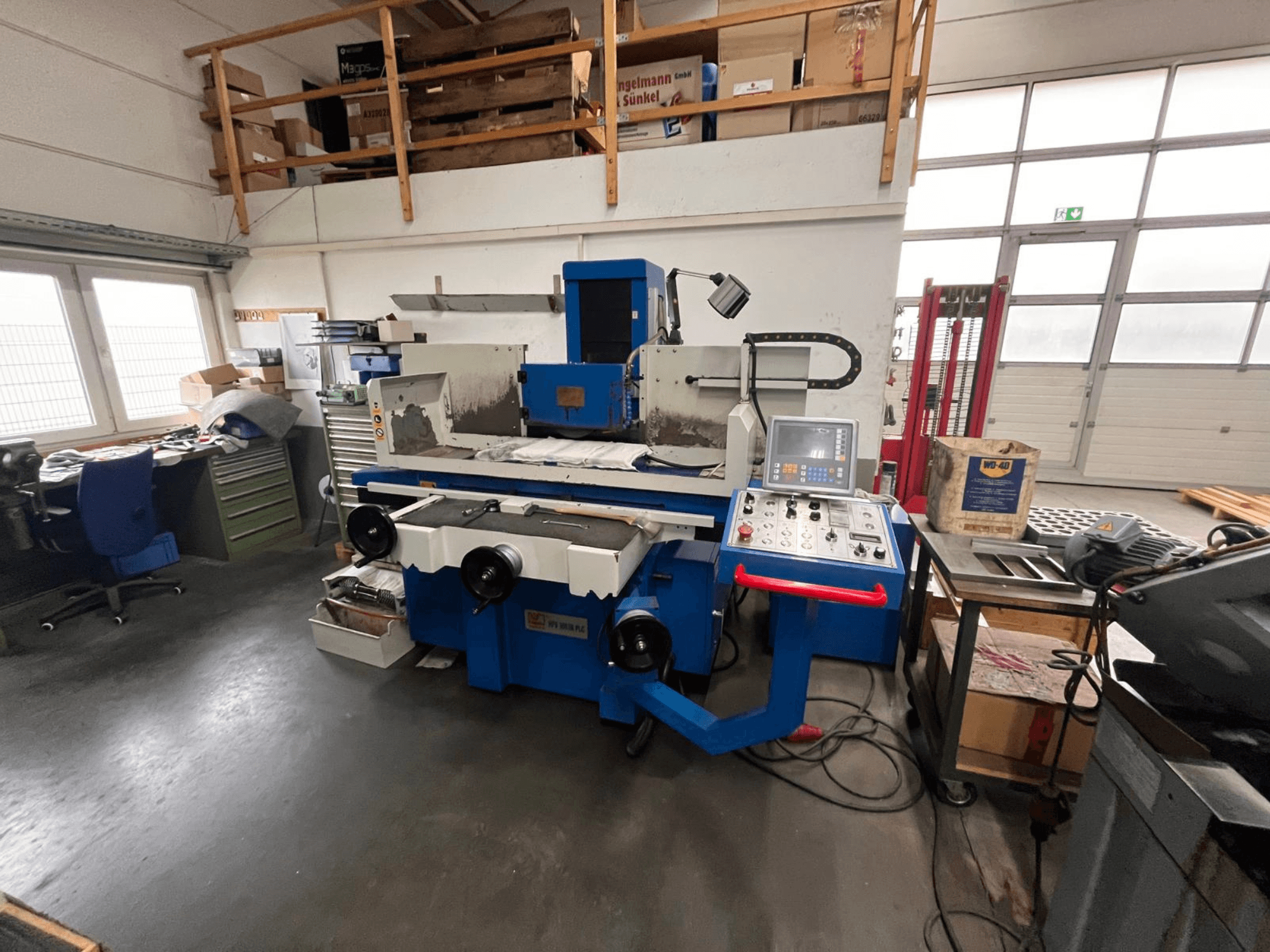 Blue surface grinder machine with digital control panel, front view, showing workspace clutter and a green tool cabinet.