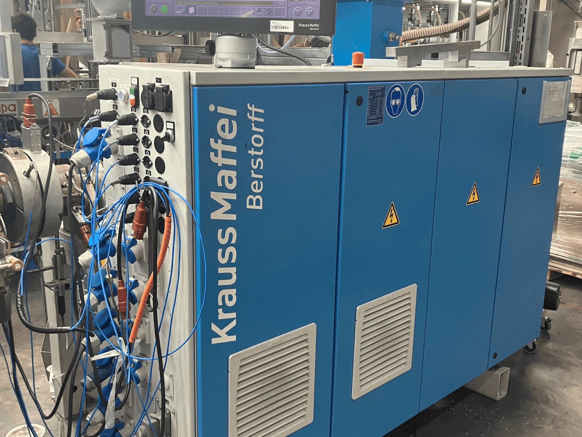 Krauss Maffei Berstorff machine in blue, front view with control panel, cables, and safety symbols. Ideal for manufacturing.