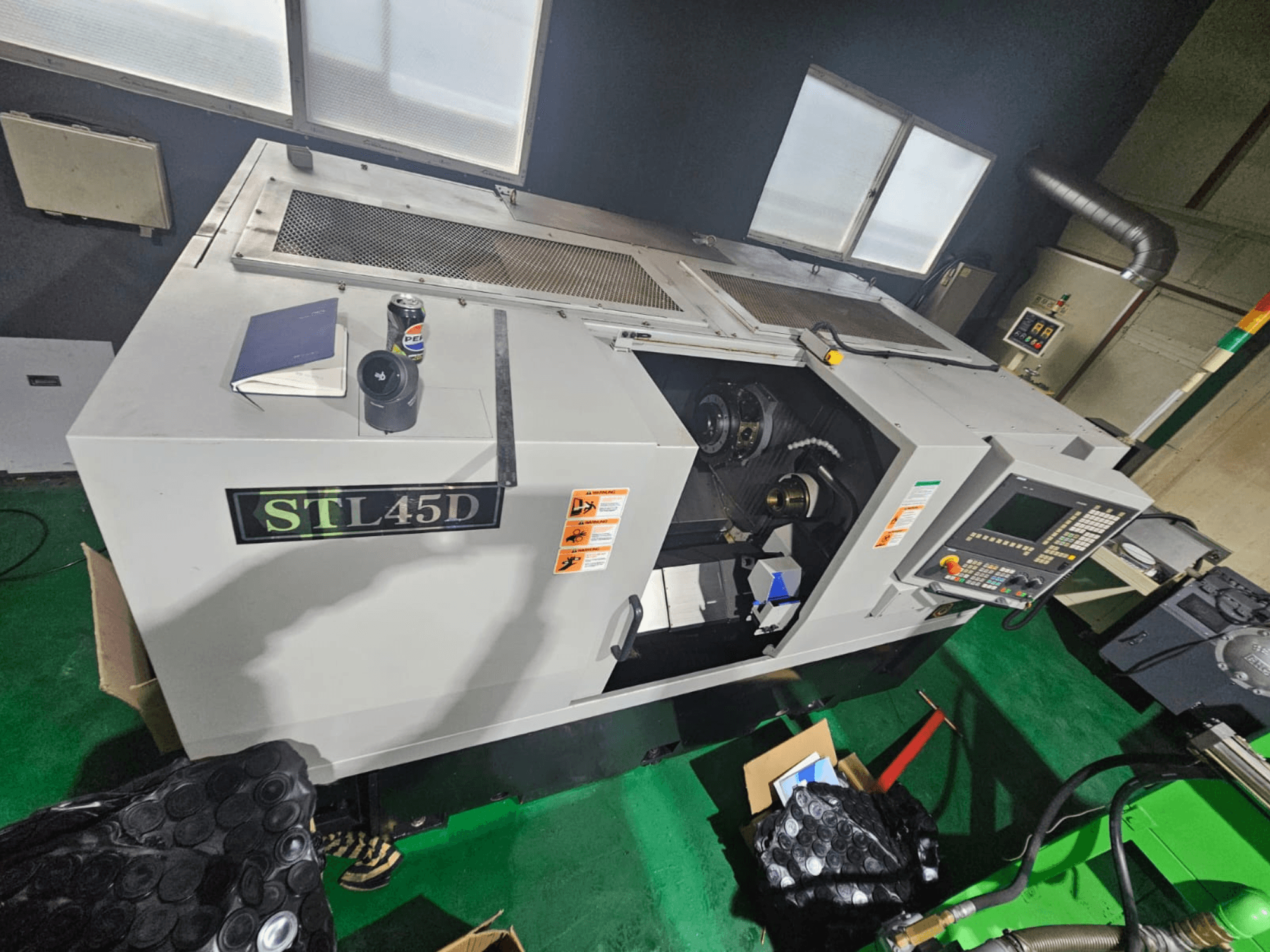STL45D CNC machine viewed from above, showing control panel, cylindrical part, notebook, and can on top.