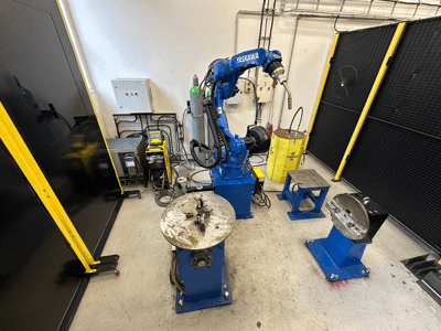 Yaskawa robotic arm in blue with two work tables, welding equipment, and safety barriers; industrial workspace front view.