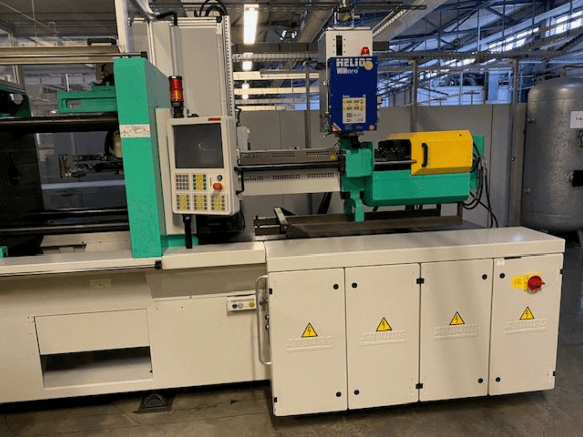 Industrial injection molding machine, model HELIO, front view, featuring control panel, safety guards, and emergency stop button.