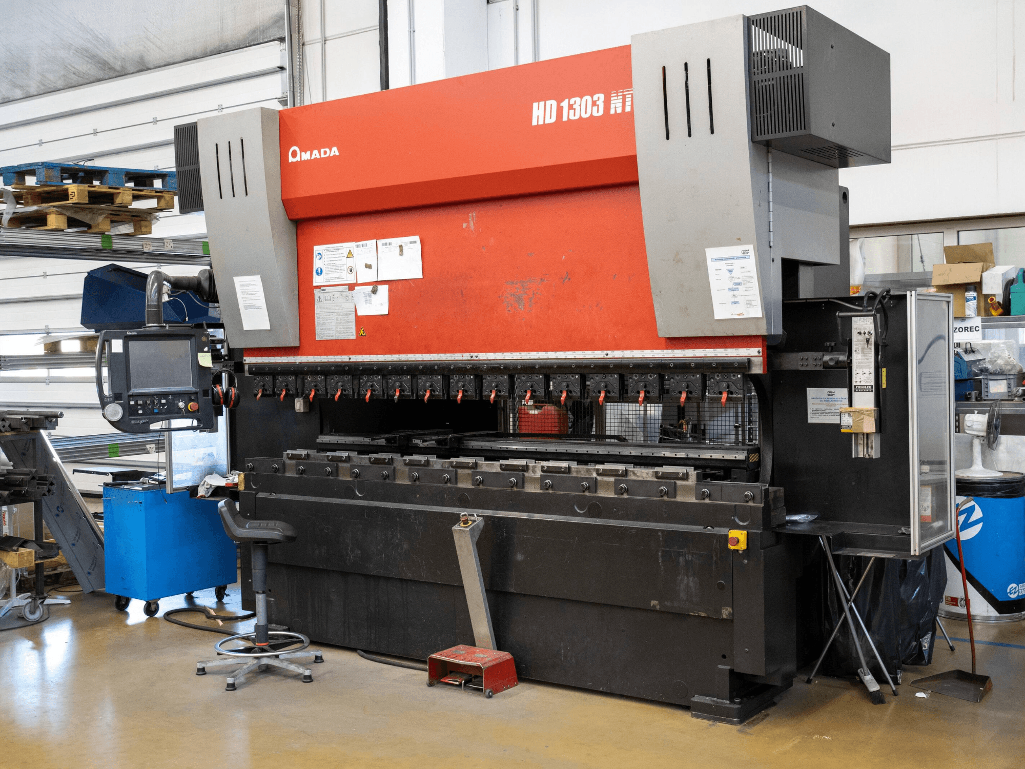 Amada HD 1303 hydraulic press brake in a workshop, front view; features multiple clamps, control panel, and adjustable stool.